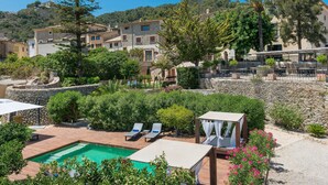 Zona de piscina con sillones de descanso, un pequeño pabellón y vistas a edificios en una colina.