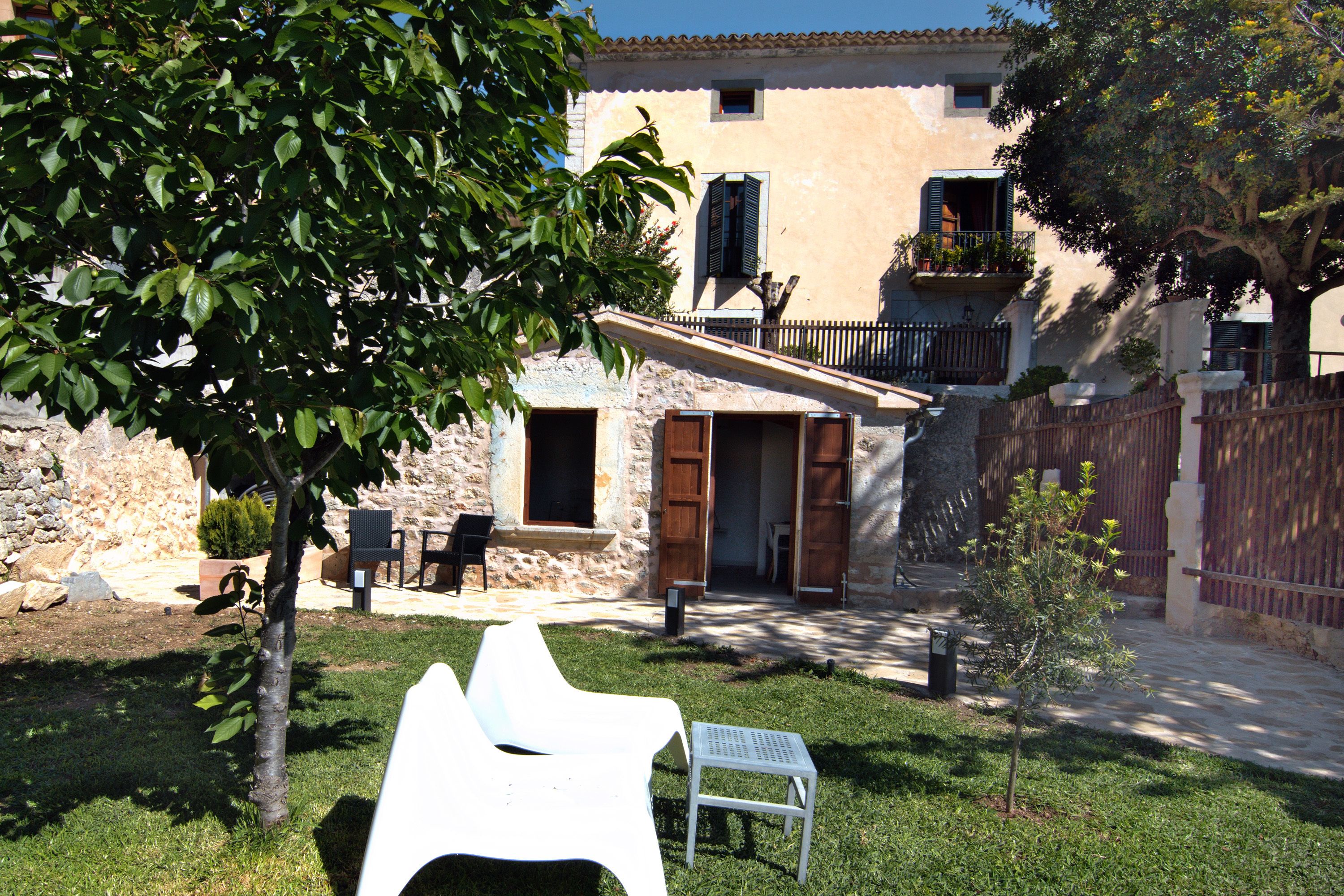 A stone building with a balcony, a small garden with a white chair and a table, and a tree in the foreground.