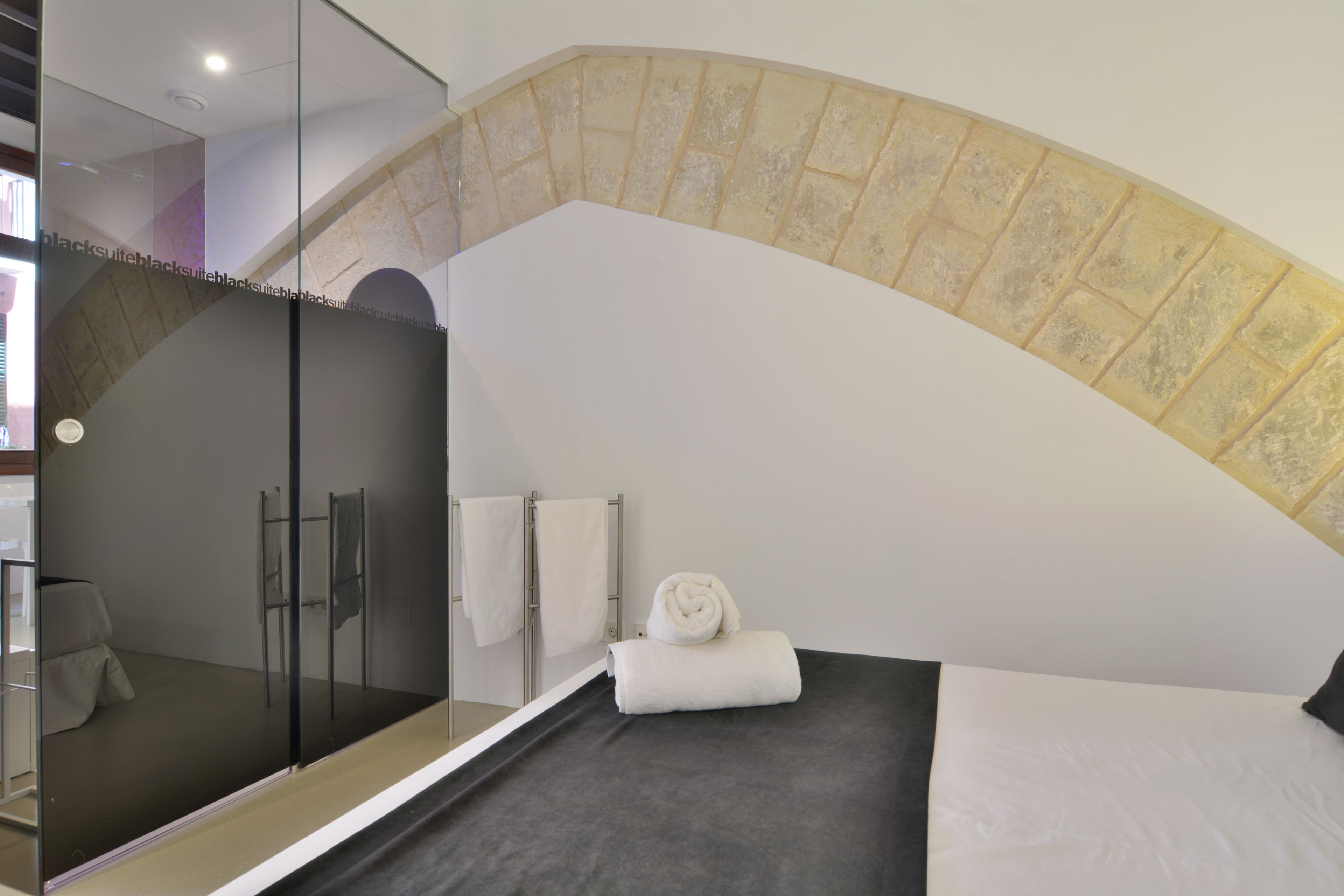 A modern hotel room with a glass-enclosed shower, a neatly made bed, and a stone archway.