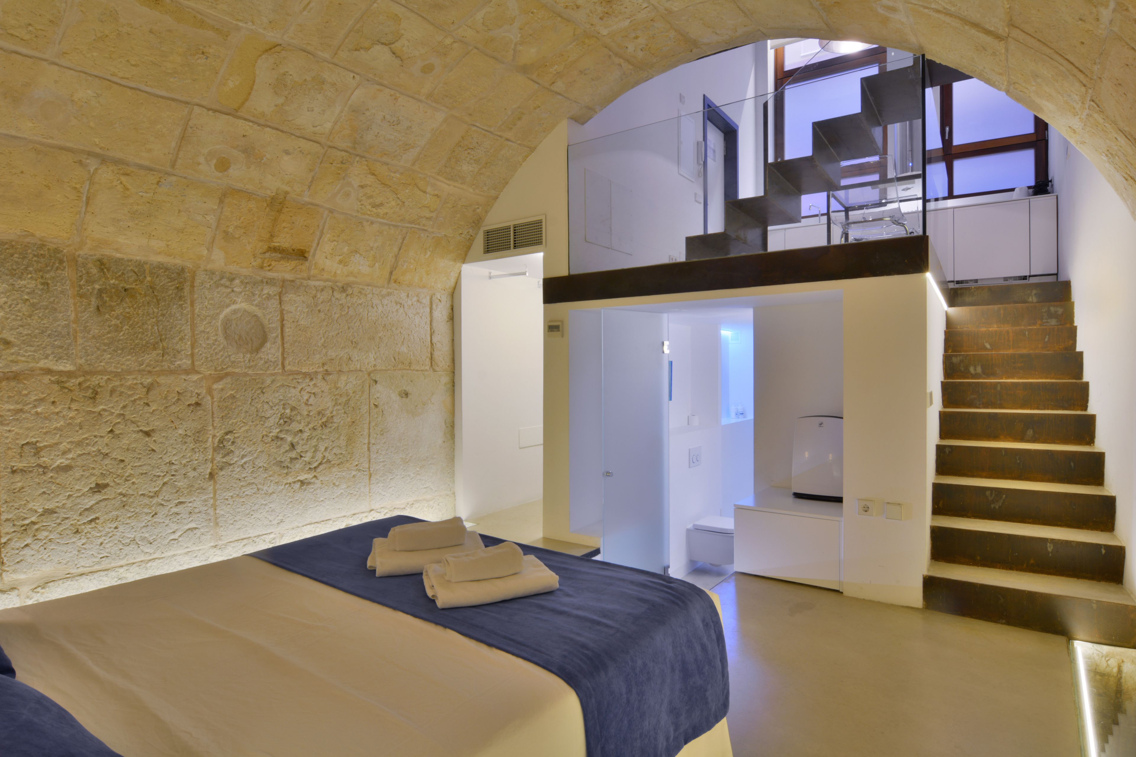 A bedroom with a stone wall, a bed with a blue headboard, a staircase leading to an upper level, and a view of the sky through an archway.