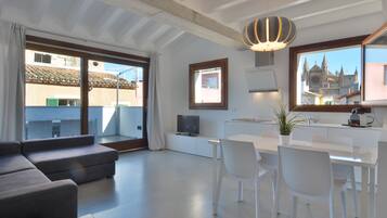 Una sala de estar moderna con un sofá, una mesa de comedor con sillas, un televisor y un ventanal con vista a un edificio.