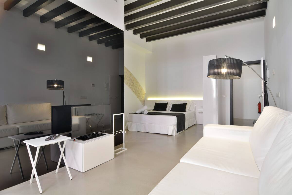 Een moderne hotelkamer met een witte bank, een klein wit tafeltje, een bed met zwart beddengoed, een grote vloerlamp en een trap naar een hoger niveau.