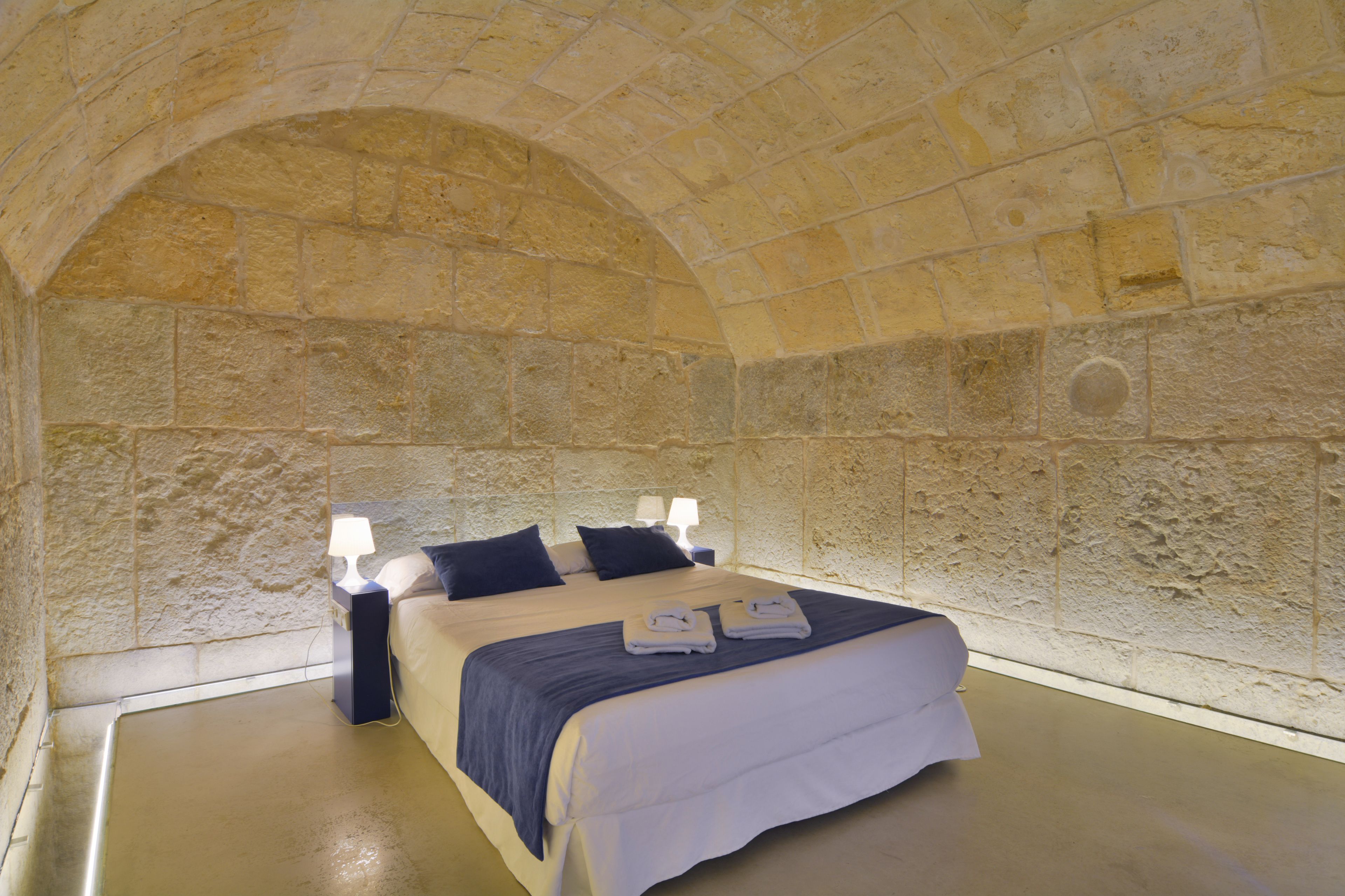 Quarto com parede de pedra, cama com colcha branca e azul, duas luminárias de cabeceira e duas toalhas dobradas na cama.