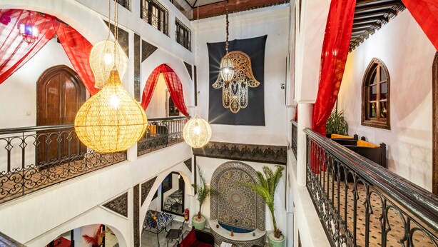 A multi-level interior with ornate decor, including a large tapestry, hanging lamps, and a balcony with a wrought iron railing. - Riad Balkisse (Marrakech)