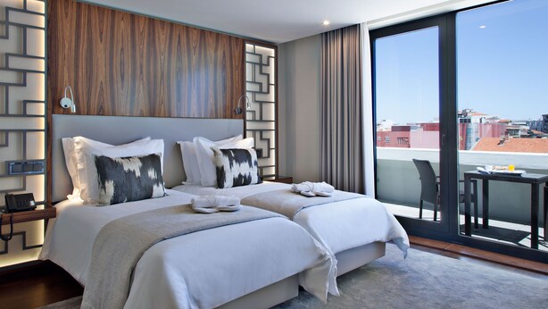 A modern hotel room with two beds, a large window, and a view of the cityscape.