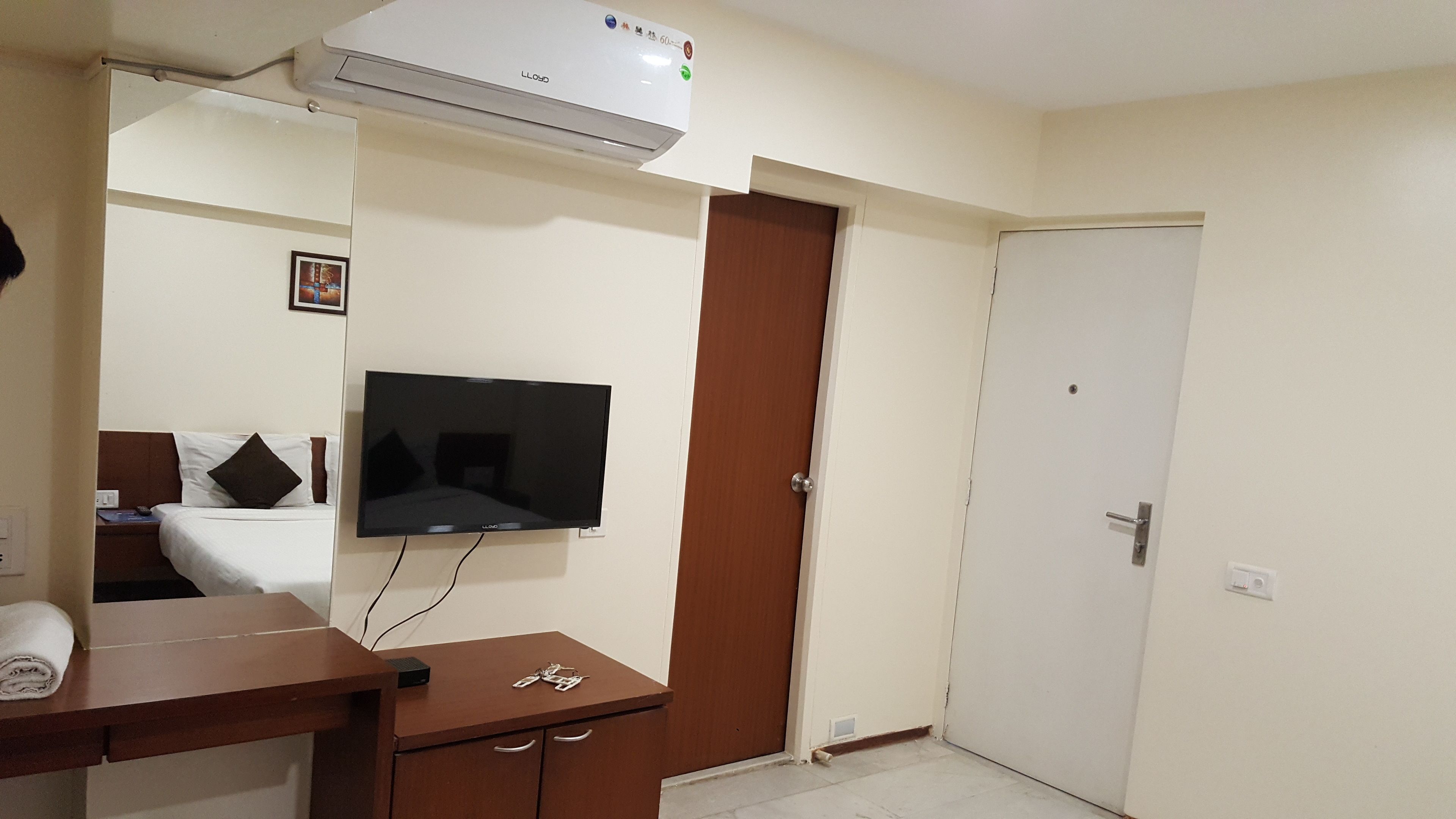 A hotel room with a bed, a TV mounted on the wall, a wooden desk, a brown door, and a white air conditioning unit.