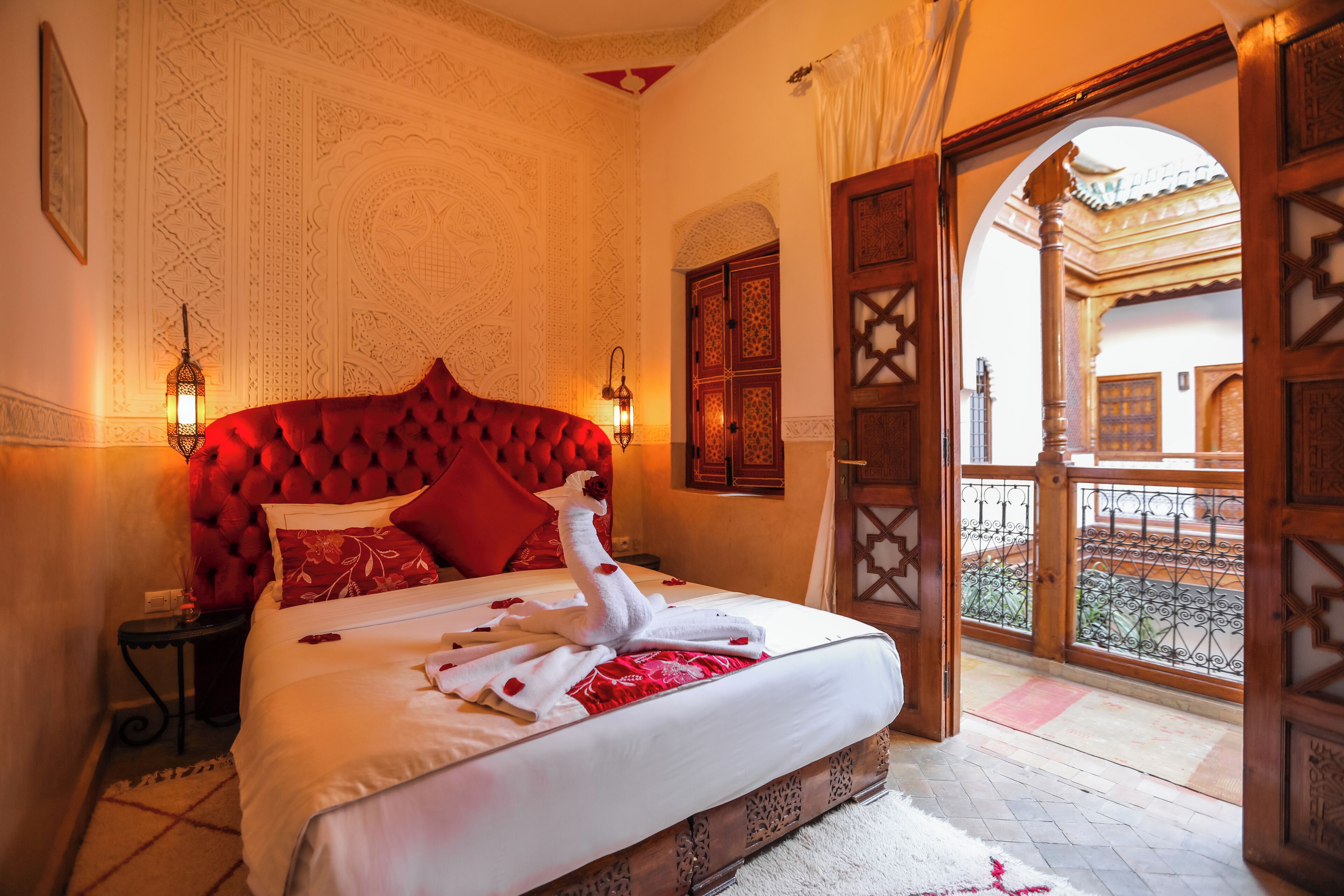 Un dormitorio con cabecera roja, cama con sábanas blancas y una toalla decorativa con forma de cisne. Hay dos lámparas fijadas a la pared y una puerta de madera que da a un área exterior.