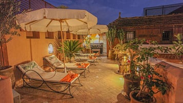 An outdoor patio area with lounge chairs and umbrellas, surrounded by potted plants and a rustic building.
