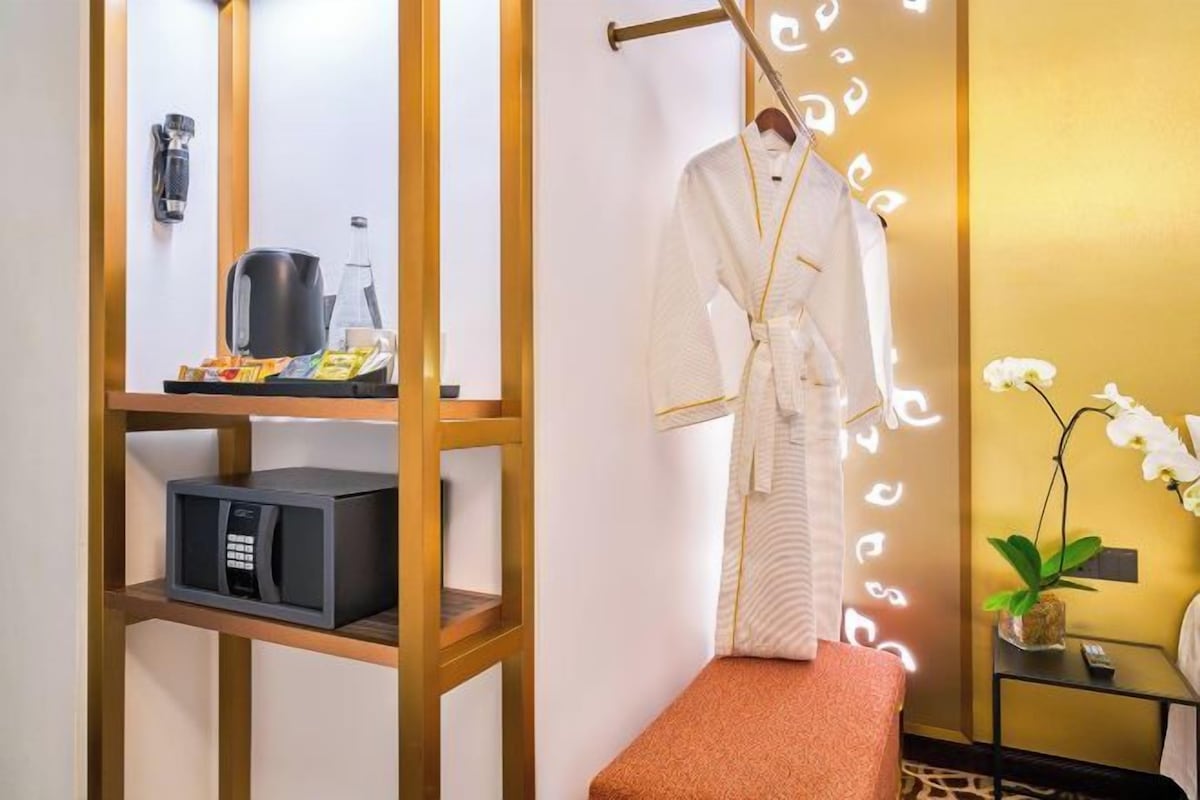Habitación de hotel con repisa de madera que sostiene una tetera, un microondas y una bandeja con aperitivos. Se observa un banco y una pequeña mesa con una planta.