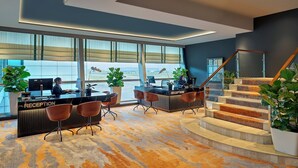 Un área de recepción moderna con un mostrador, dos recepcionistas y una escalera que sube a un piso superior.