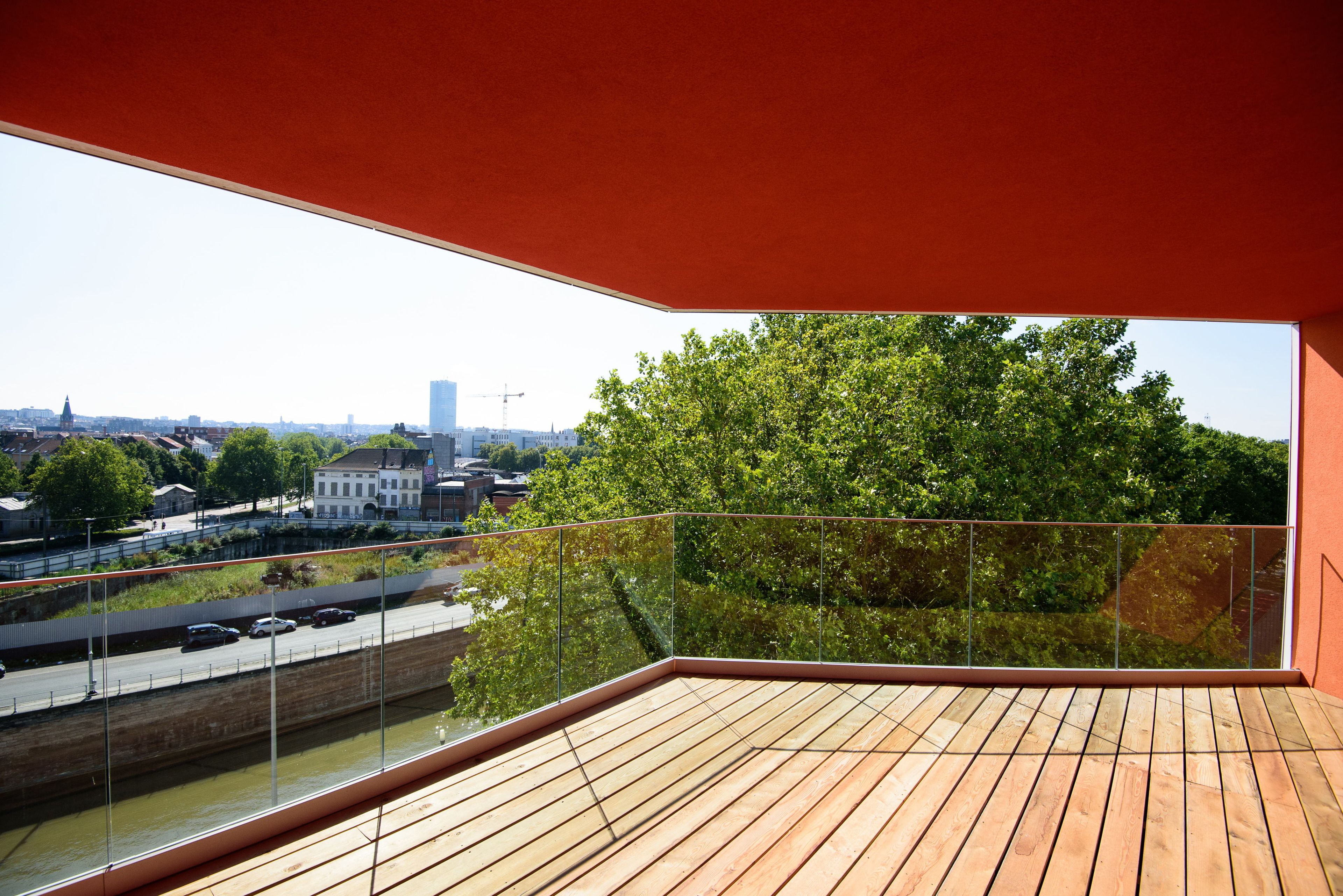 A balcony with a wooden floor and glass railing, overlooking a river with a bridge and buildings in the background.