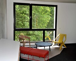 Ein Zimmer mit einem großen Fenster, einem Holzstuhl, einem gelben Stuhl, einer roten Bank und einem kleinen runden Tisch.