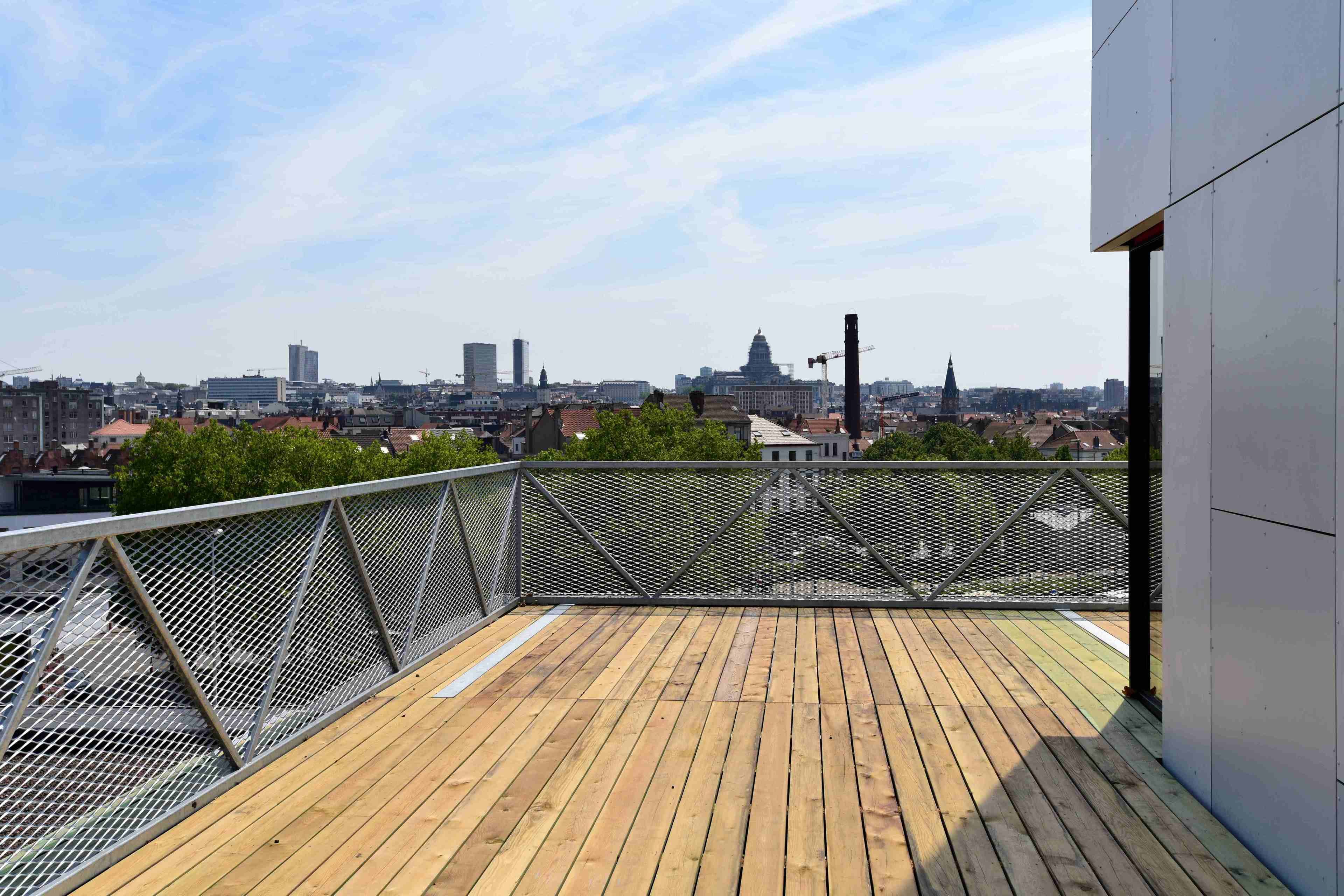 Terraza en la azotea con cubierta de madera y barandilla de malla metálica, con vistas a un paisaje urbano que incluye varios edificios y una aguja de iglesia.