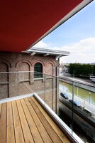 En træterrasse med rødt markise, der giver udsigt over en murstensbygning med buevinduer og en kanal med både.
