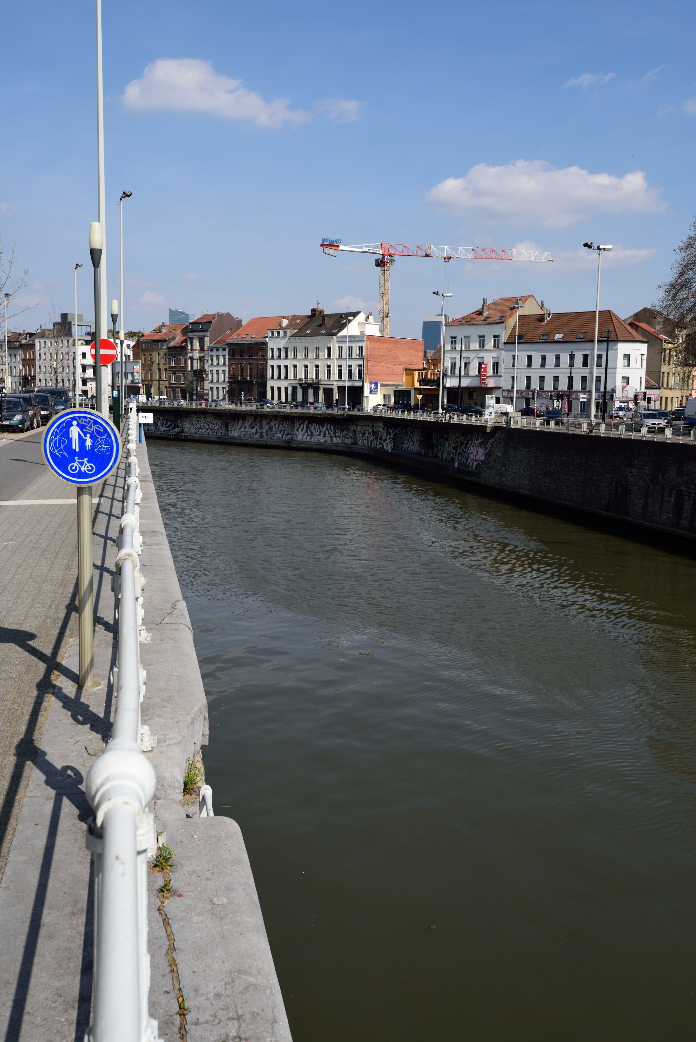 A riverside walkway with a blue sign indicating a pedestrian and bicycle path, a construction crane in the background, and buildings along the riverbank.