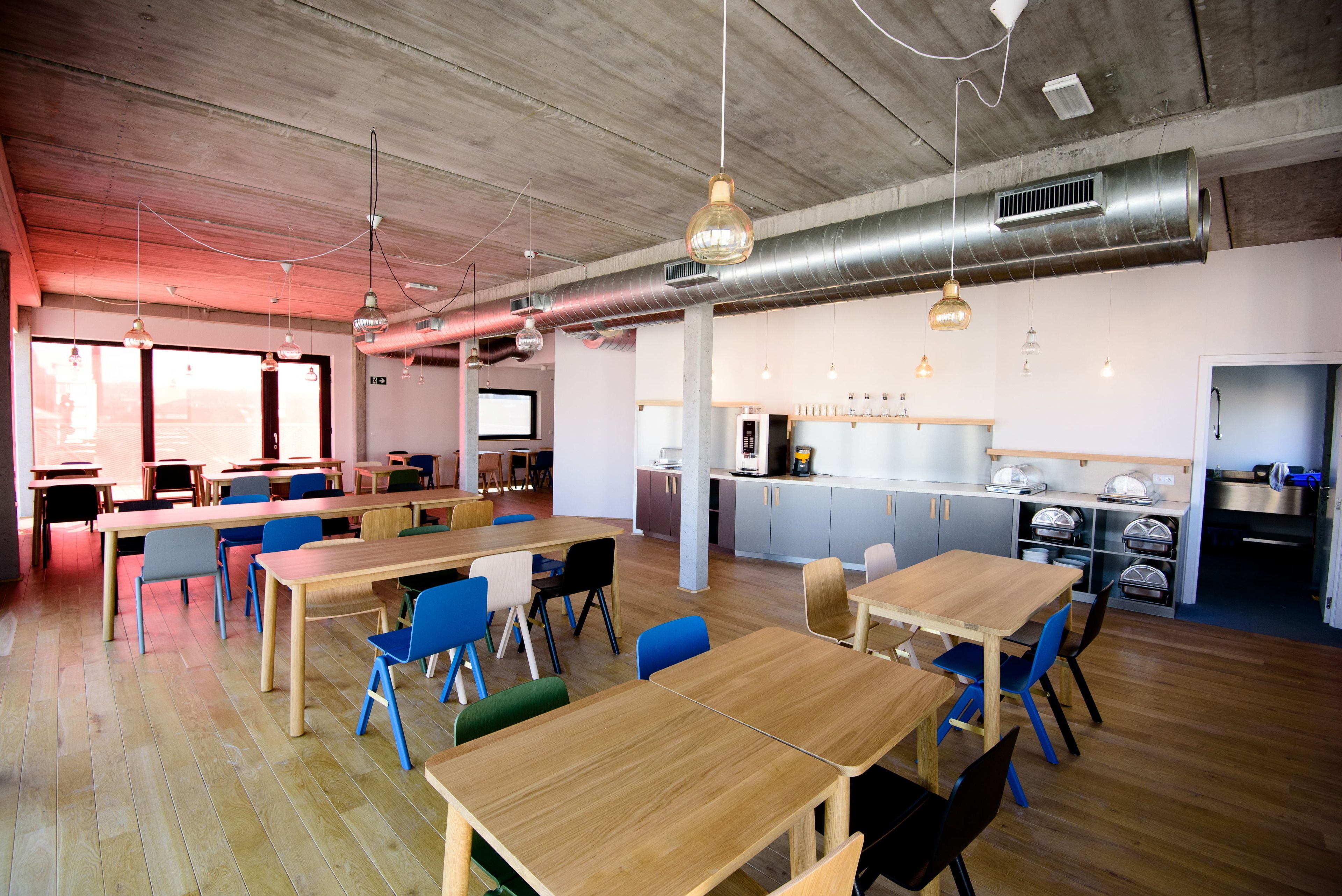 Un vaste espace de salle à manger avec des tables et des chaises en bois, un coin cuisine équipé d’appareils électroménagers en acier inoxydable et une grande fenêtre qui laisse entrer la lumière naturelle.