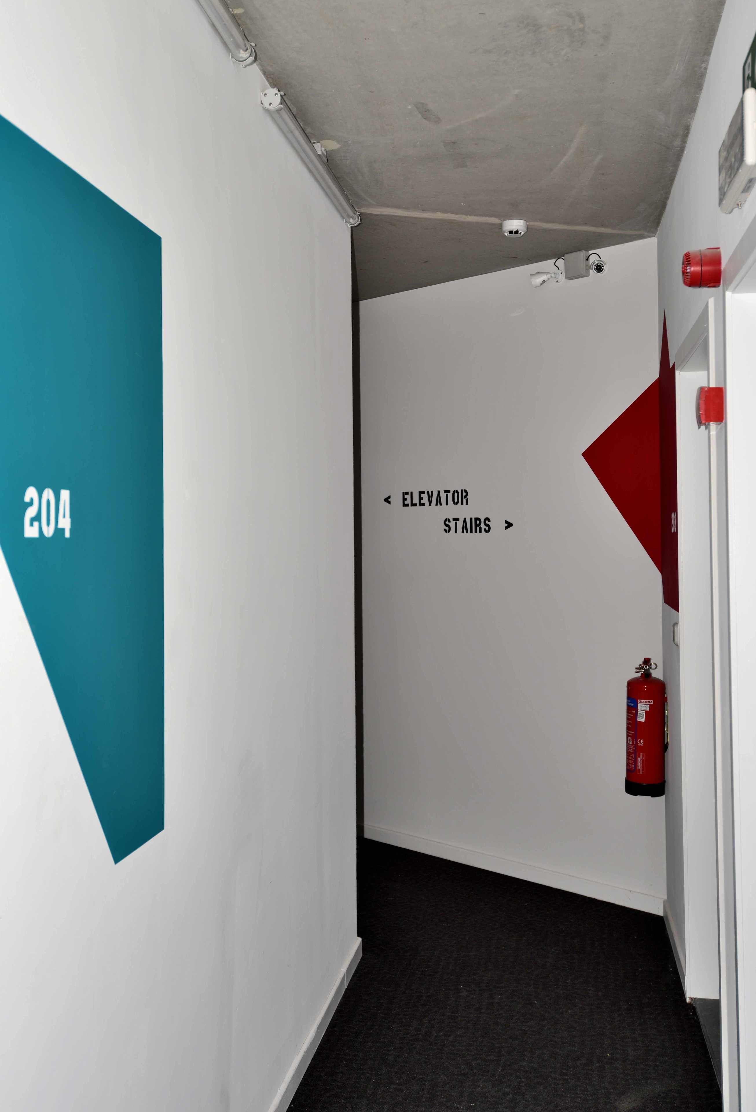 Un couloir avec un mur de couleur turquoise sur la gauche, un mur blanc sur lequel on peut lire les mots « Escalier d’accès à l’ascenseur », et un extincteur rouge sur la droite.