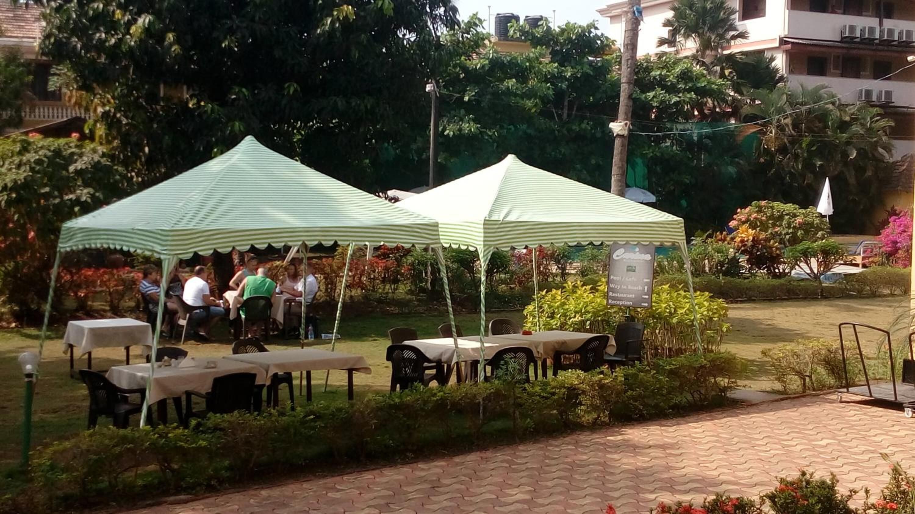 An outdoor seating area with tables and chairs under striped canopies, surrounded by greenery and a paved walkway.