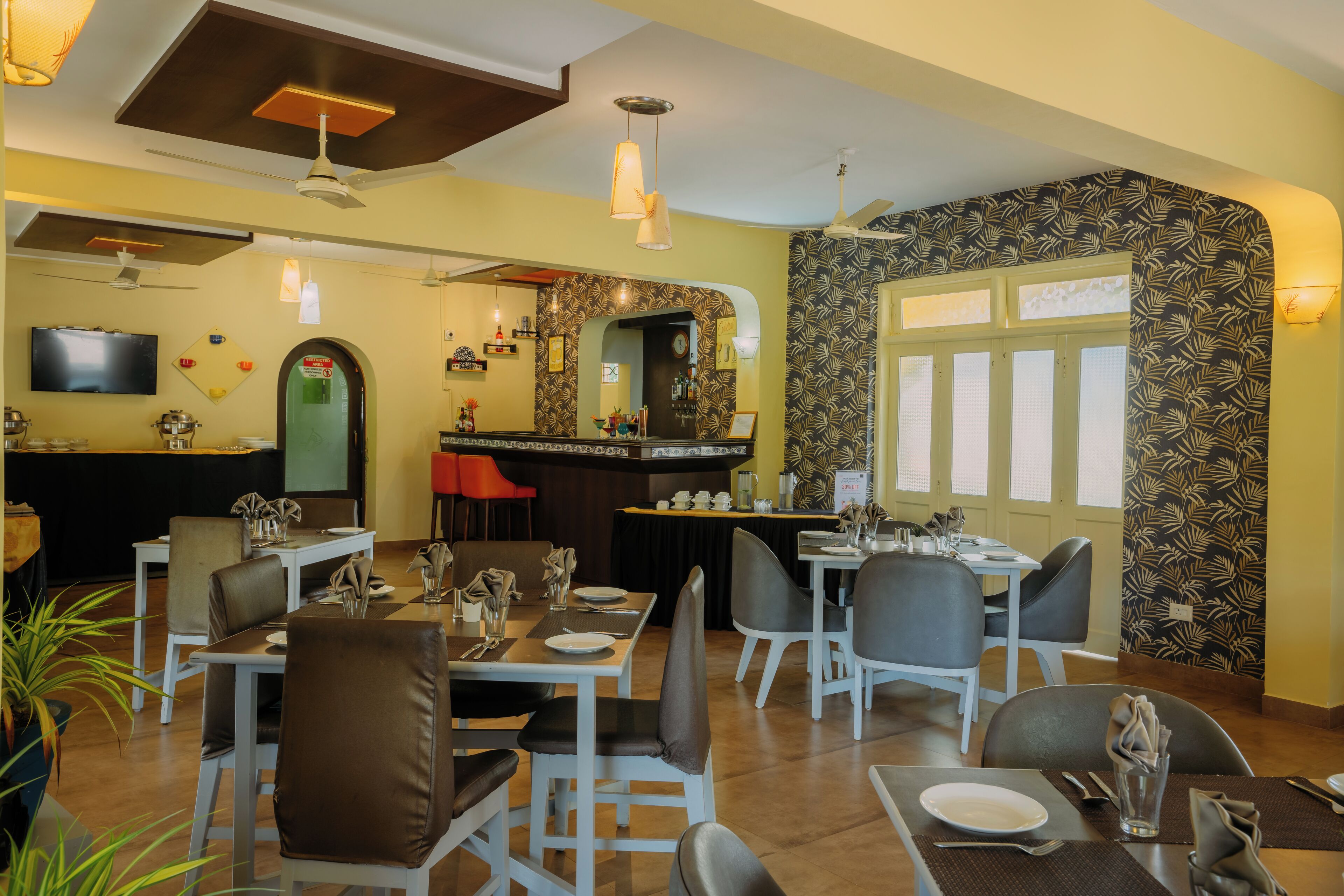 A restaurant with multiple dining tables and chairs, a bar area, and a television mounted on the wall.