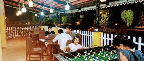 An indoor recreational area with a foosball table, tables, chairs, hanging lights, and a white picket fence.