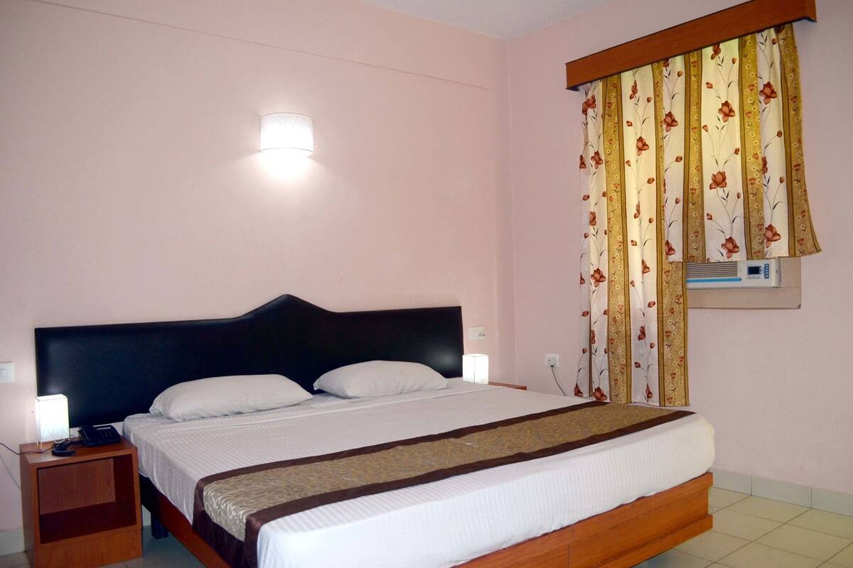 A bedroom with a bed, bedside lamps, a window with curtains, and a wall-mounted air conditioning unit.