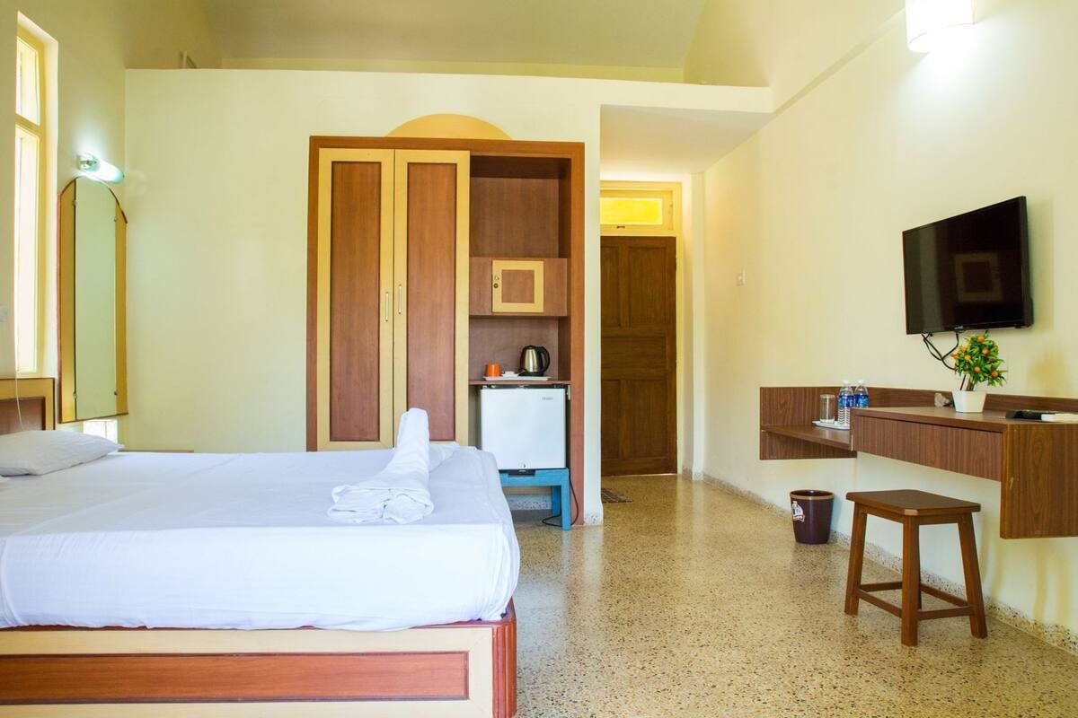 A hotel room with a bed, a desk with a mirror, a television, and a kitchenette.