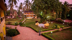 A well-maintained garden with a paved walkway, palm trees, and a building with a balcony in the background. - Carina Beach Resort (Benaulim)