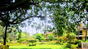 An outdoor dining area with a table set for two, surrounded by lush greenery and a view of a yellow building. - Carina Beach Resort (Benaulim)