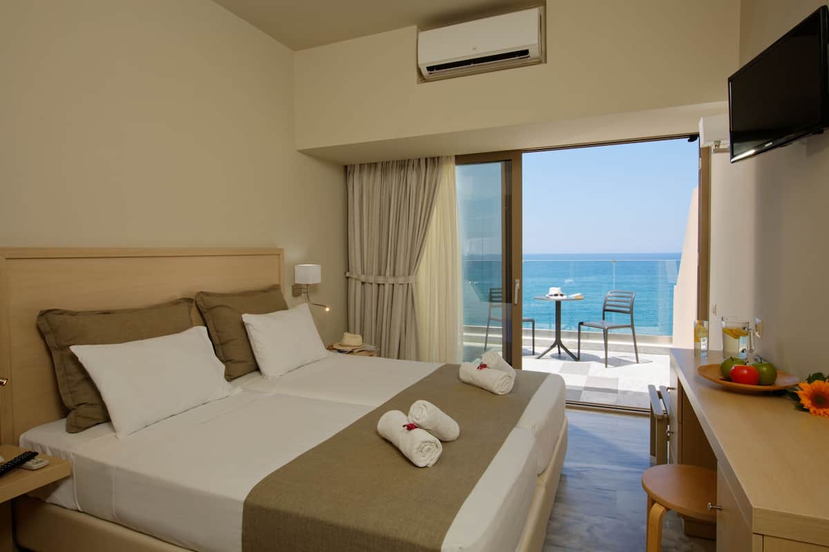 Une chambre d’hôtel avec un grand lit, une télévision, un balcon avec vue sur la mer et un coin petit-déjeuner avec des fruits et une fleur.