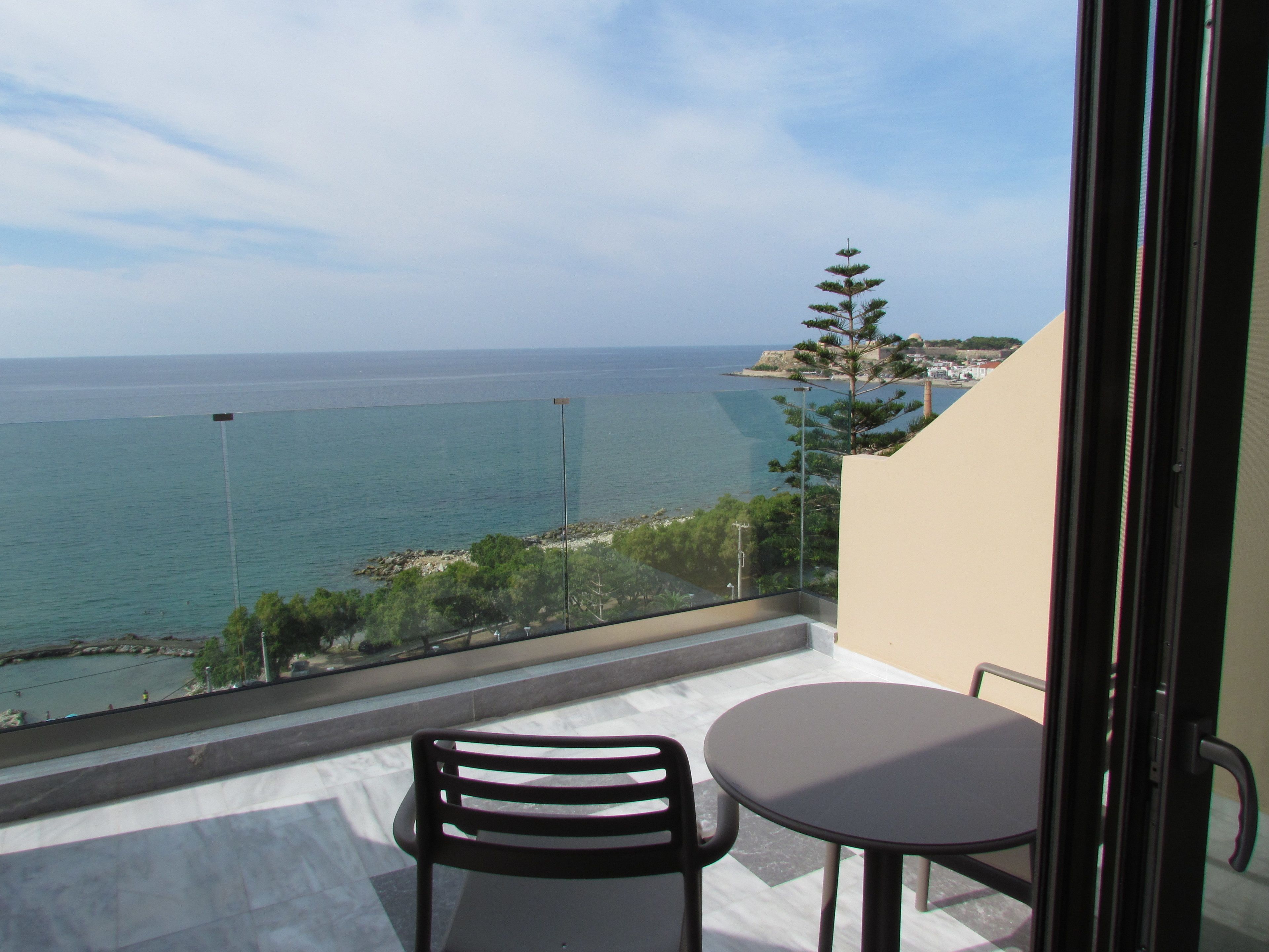 A balcony with a table and two chairs, overlooking a coastal view with the sea and a distant shoreline.