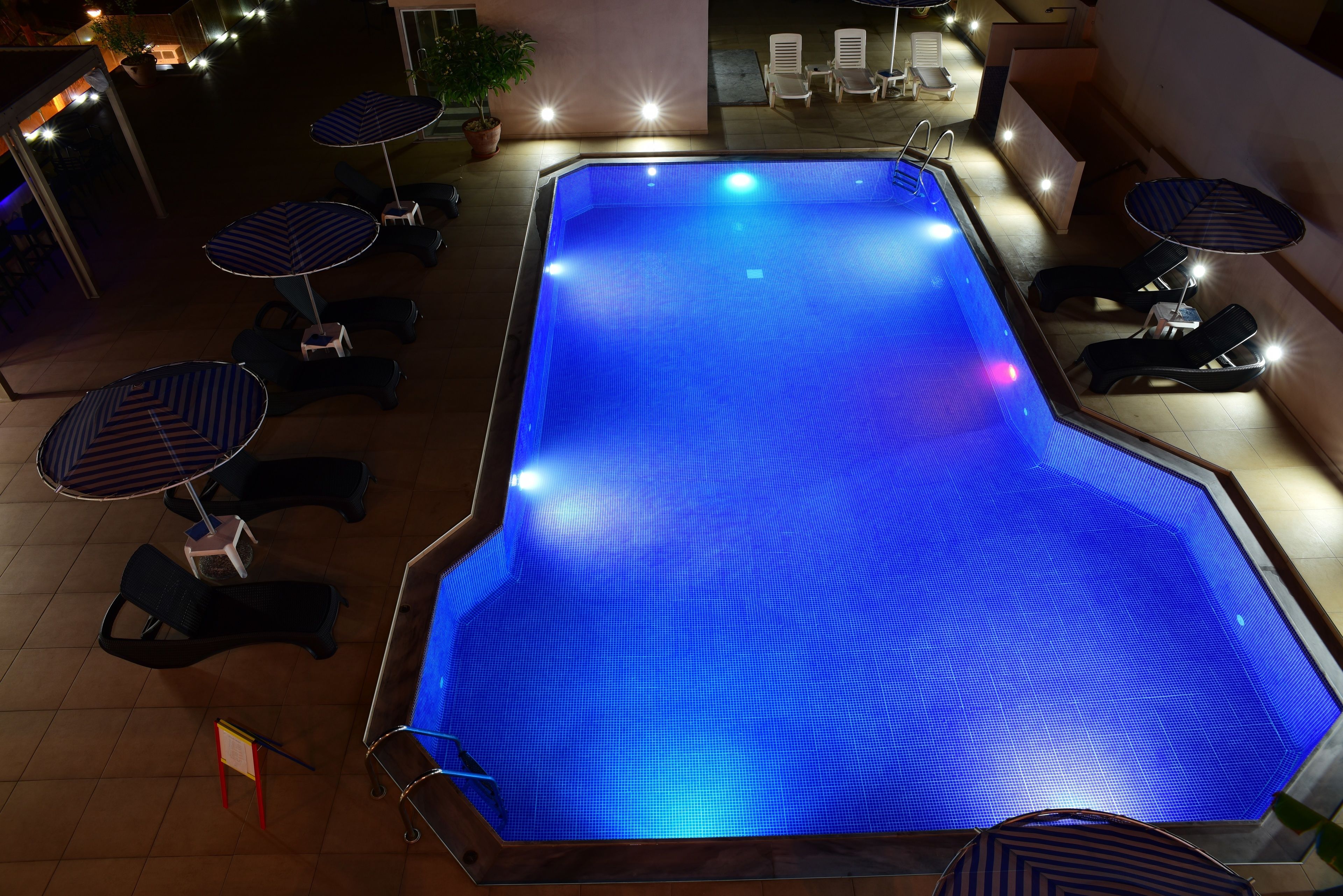 Une piscine entourée d’une terrasse et de murs recouverts de carrelage, avec des chaises longues et des parasols à proximité.