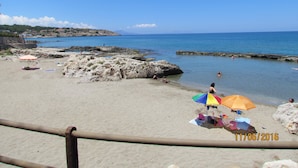 En strand med klippepartier, klart blåt vand og et par personer.