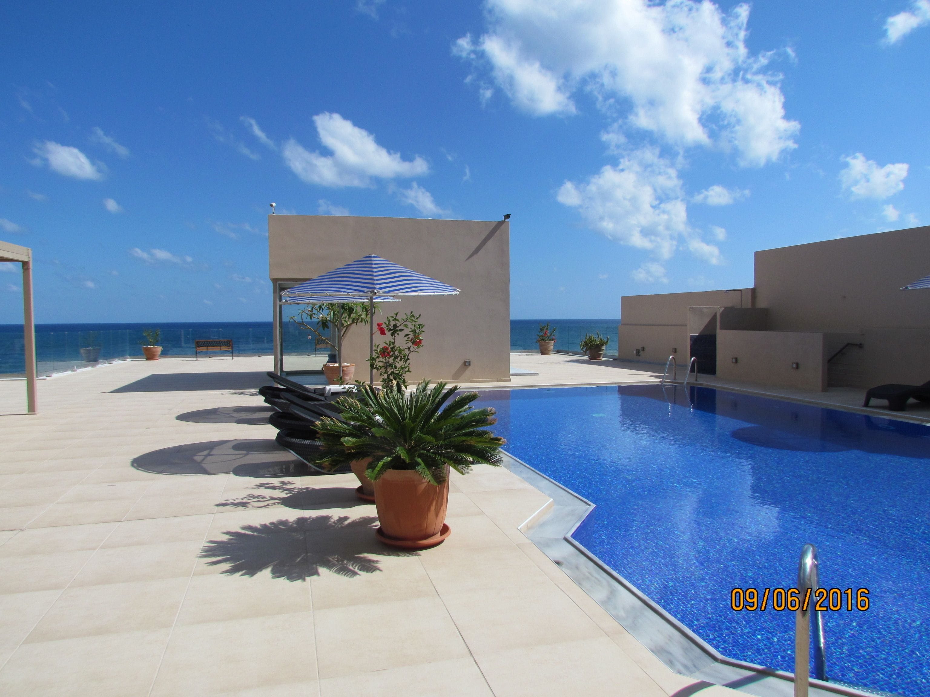 Zona junto a la piscina con vista despejada al océano, una sombrilla azul y blanca, y una planta en maceta.