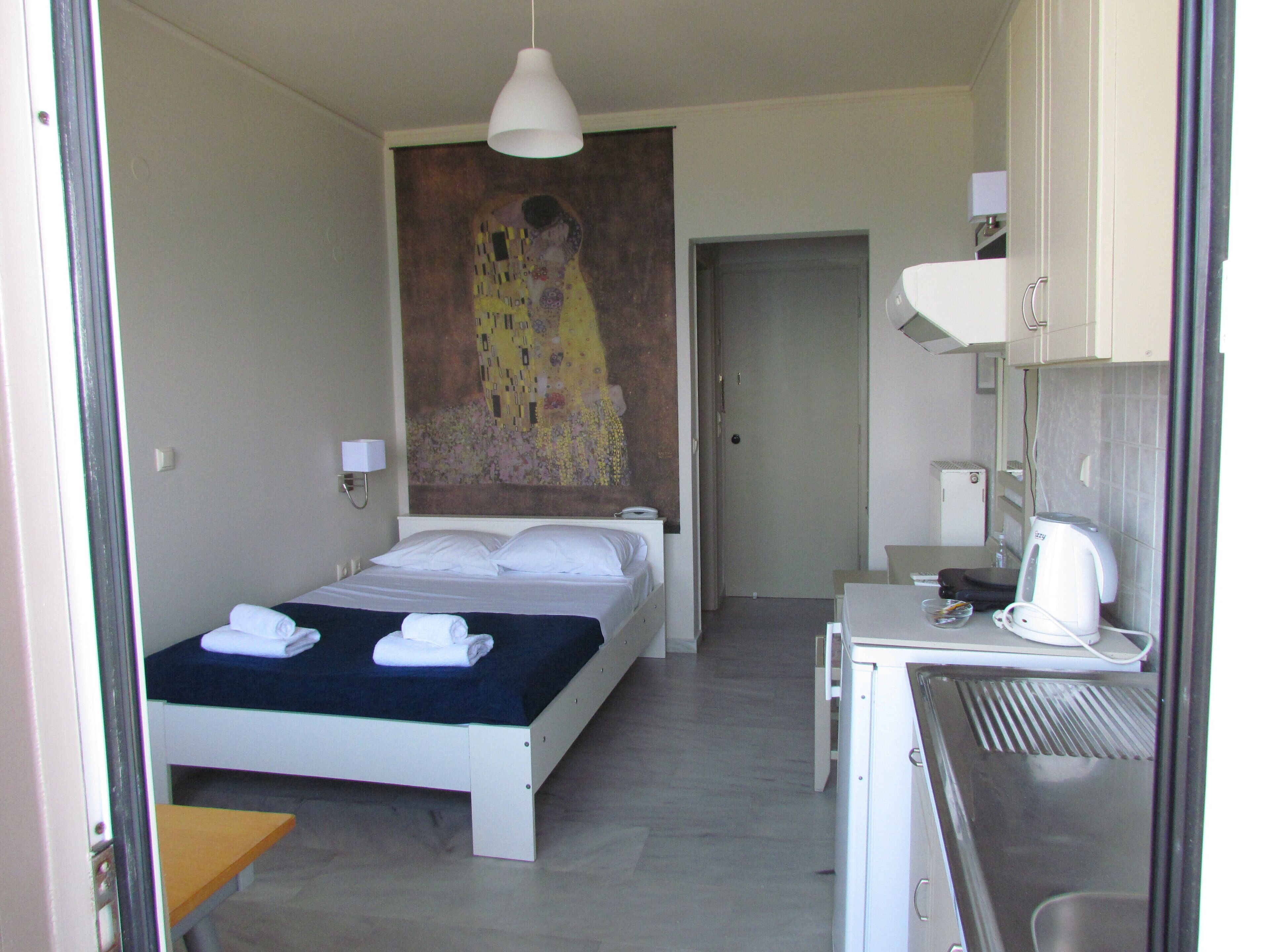 A hotel room with a bed, a desk, a kettle, and a painting on the wall.