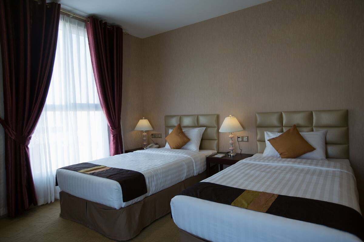 A hotel room with two beds, each with a headboard, bedside tables with lamps, and a window with curtains.