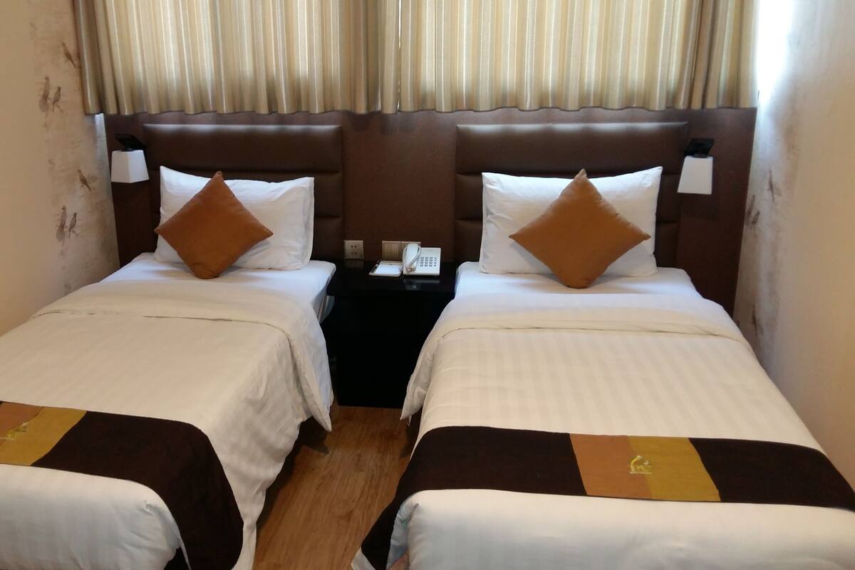 A hotel room with two single beds, each with a brown headboard, white bedding, and a brown accent pillow. There is a bedside table with a phone and a lamp.