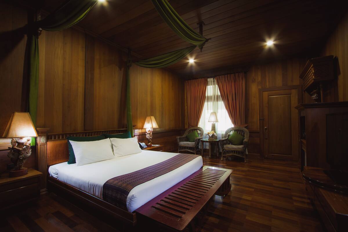Ein Hotelzimmer mit einem großen Bett, zwei Nachttischen mit Lampen, einer hölzernen Bank, einem Sessel, einem kleinen Tisch mit Lampe und einem Kleiderschrank.