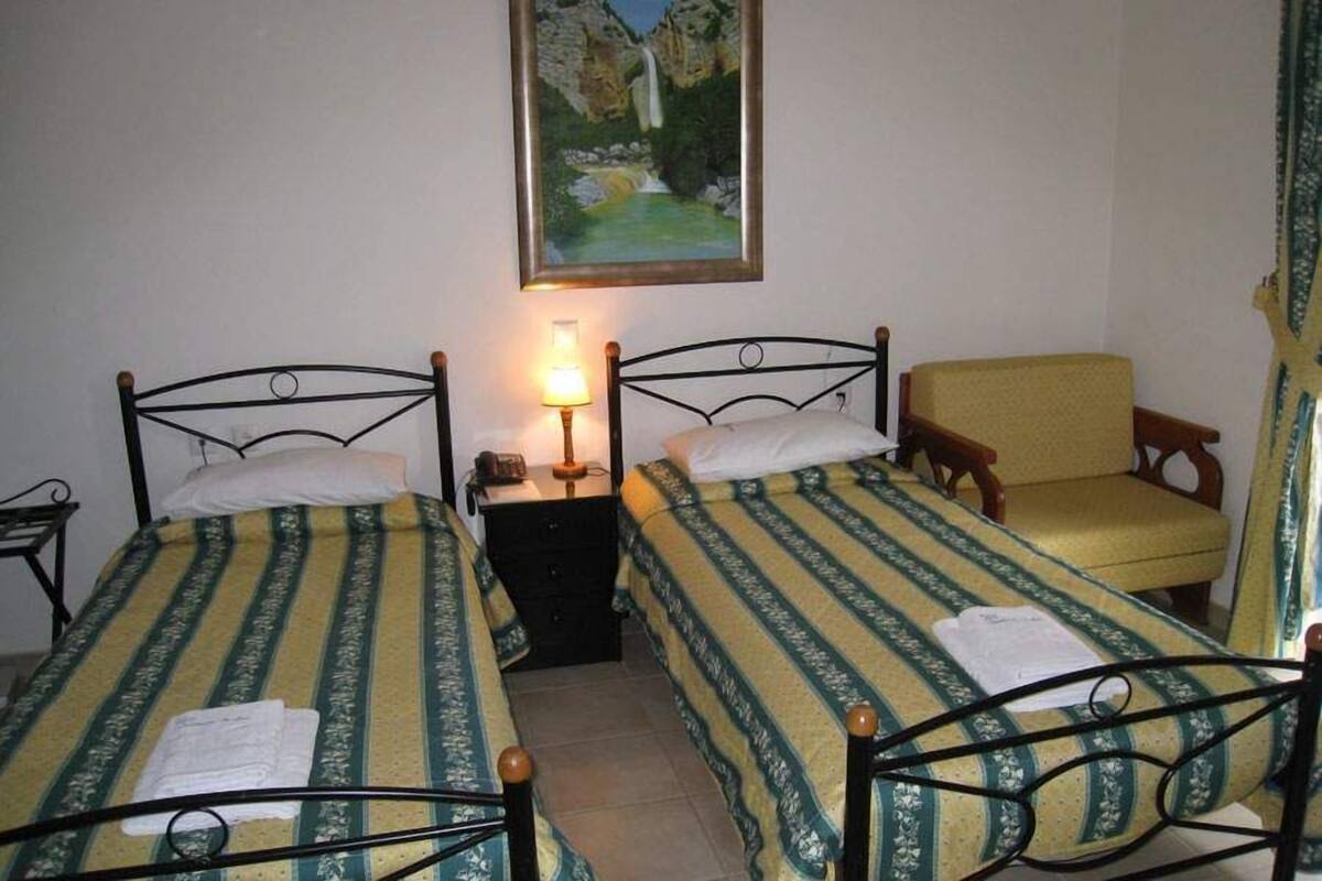 A bedroom with two single beds, a nightstand with a lamp, a chair, and a painting on the wall.