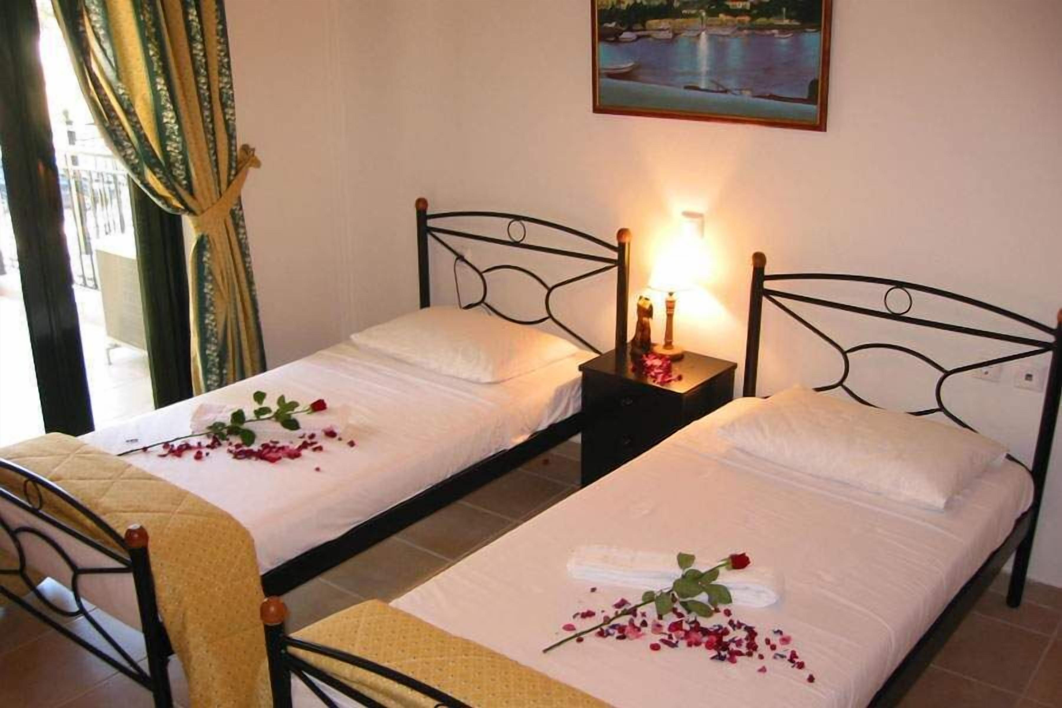 Habitación de hotel con dos camas individuales, cada una con una colcha blanca y una rosa decorativa. Hay dos mesitas de noche con velas y un cuadro sobre las camas.