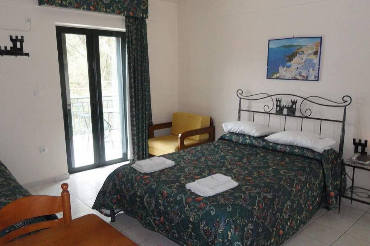 A bedroom with a bed, a chair, a table, a painting, and a window with curtains.