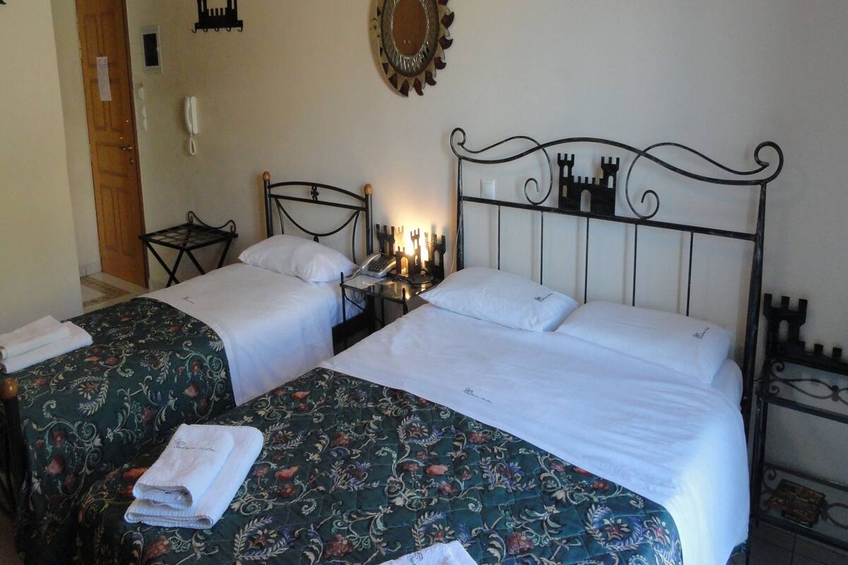 A hotel room with two beds, a wooden door, a wall clock, and a small table with a telephone.