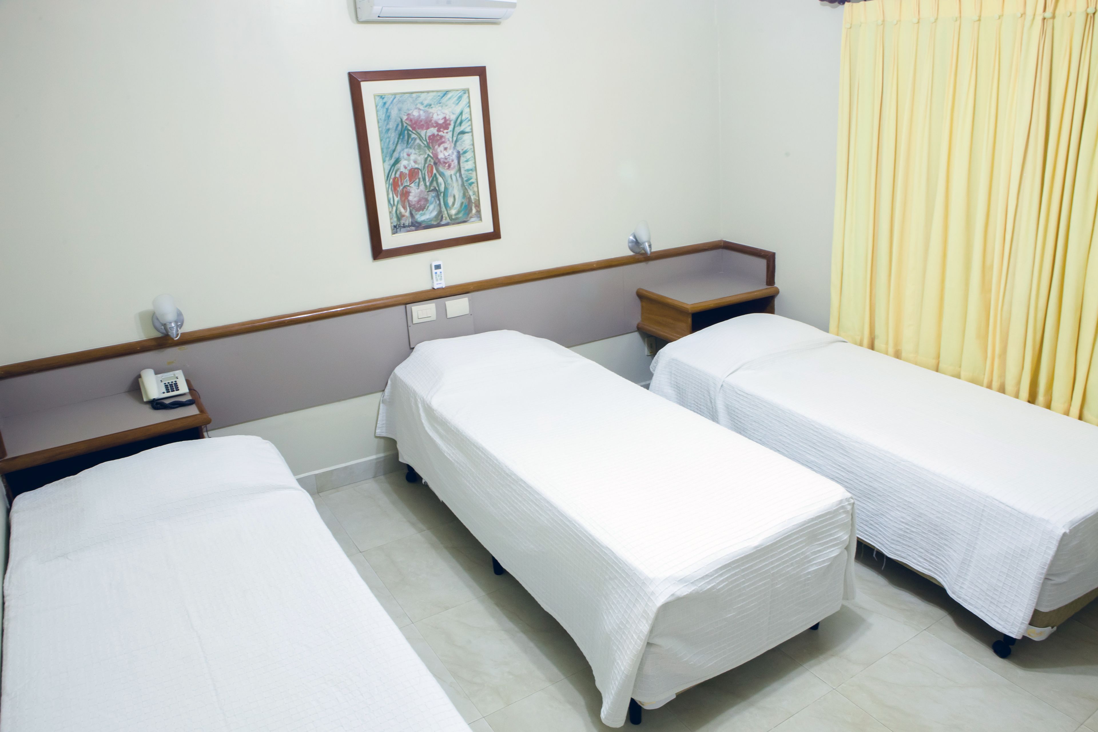 A hotel room with three single beds, a bedside table with a phone, a framed artwork on the wall, and a window with yellow curtains.