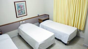 Quarto de hotel com duas camas de solteiro, uma mesinha de cabeceira, uma luminária e um quadro na parede, com cortina amarela.