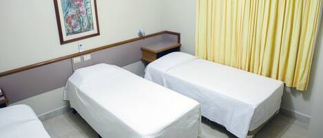 Habitación de hotel con dos camas individuales, una mesita de noche, una lámpara de noche, un cuadro en la pared y una cortina amarilla.