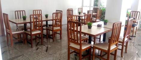 Comedor con mesas y sillas de madera, plantas en macetas y amplios ventanales que dejan entrar luz natural.