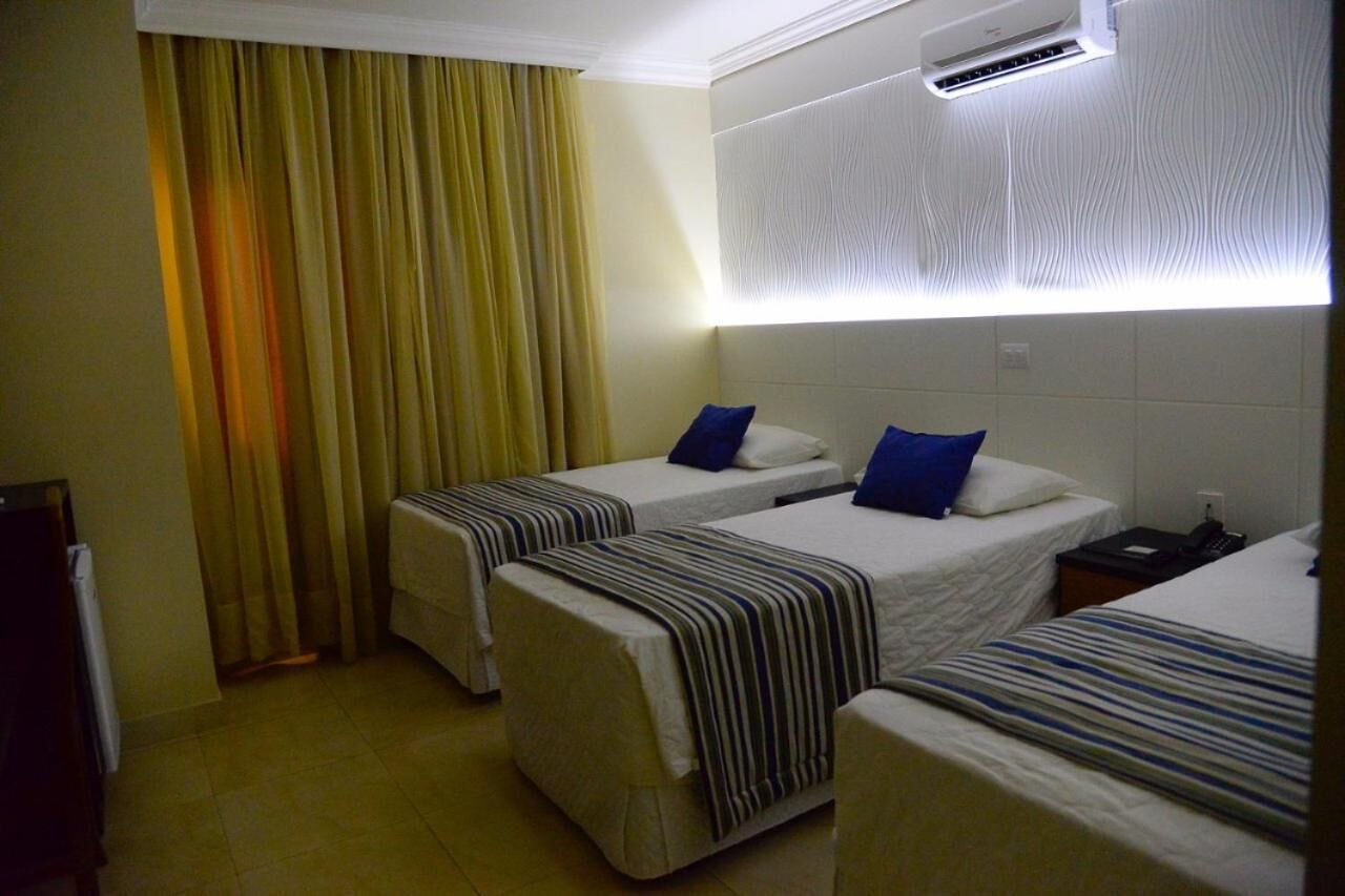 Habitación de hotel con dos camas individuales, una mesita de noche con lámpara y una ventana con cortinas.