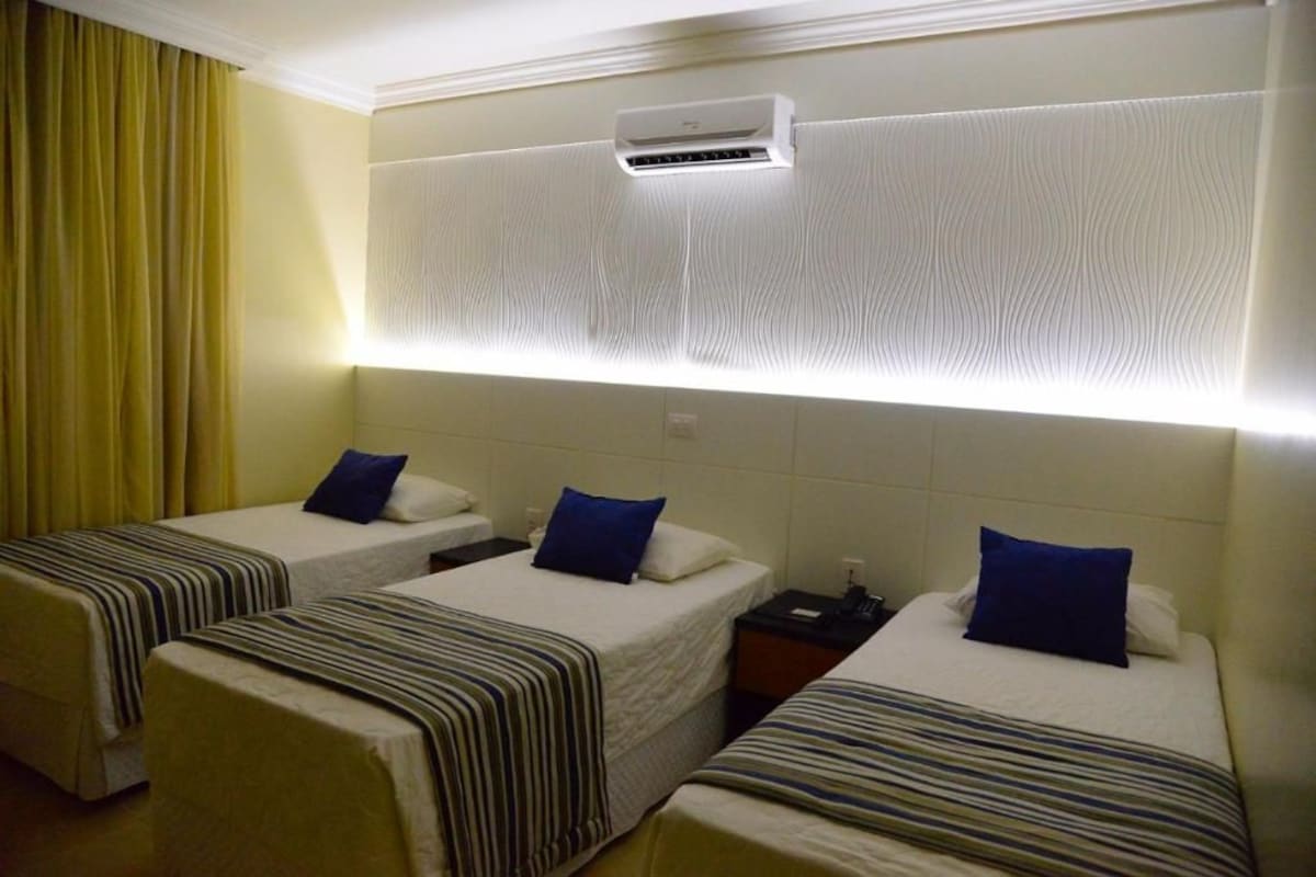 A hotel room with two single beds, each with a striped bedspread and a blue pillow. There is a wall-mounted air conditioning unit above the beds.