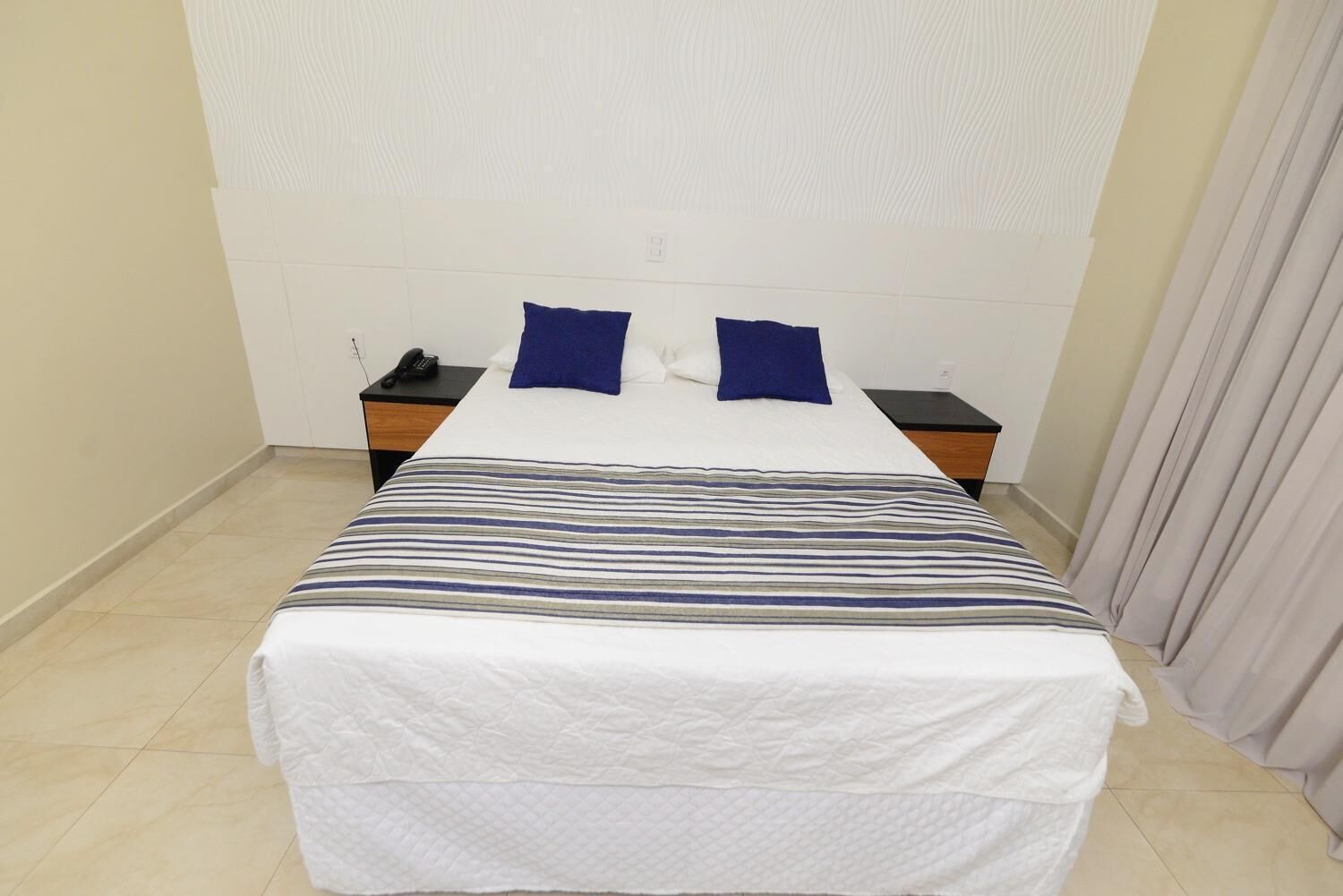 Un letto ben rifatto con copripiumino a righe e due cuscini, un comodino con un telefono e una tenda sulla destra.