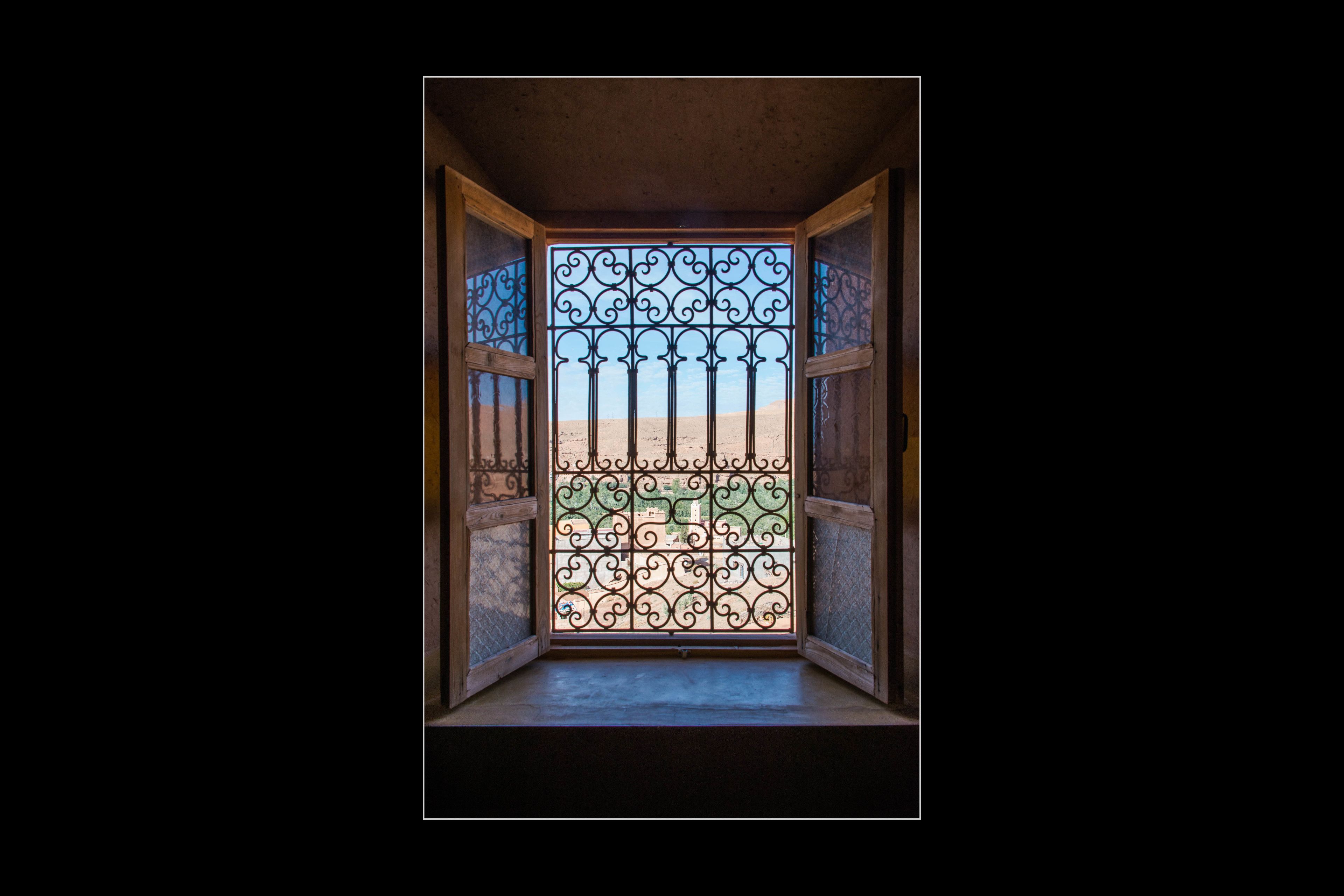 A view through a barred window with a metal gate, leading to a sandy area with a building in the background.