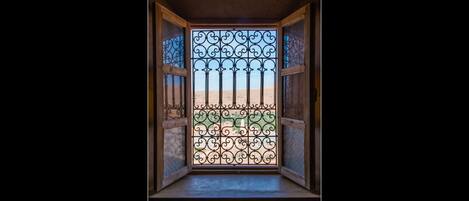 A view through a barred window with a metal gate, leading to a sandy area with a building in the background.